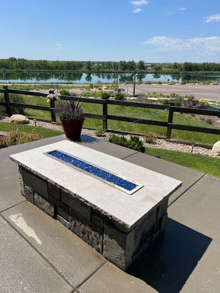Patio with paving stones, seating, and a tan umbrella next to a house with windows and green landscaping.