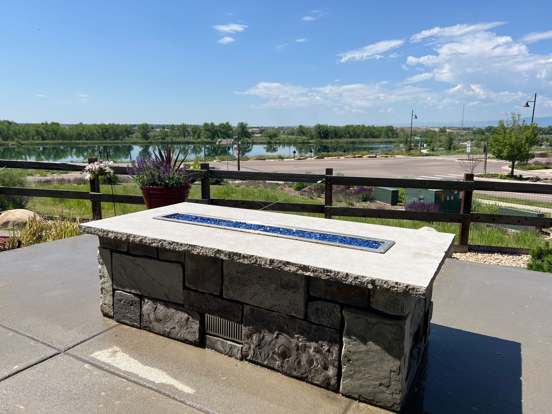 Stone fire pit with blue glass on a patio overlooking a lake and fence under a blue sky.