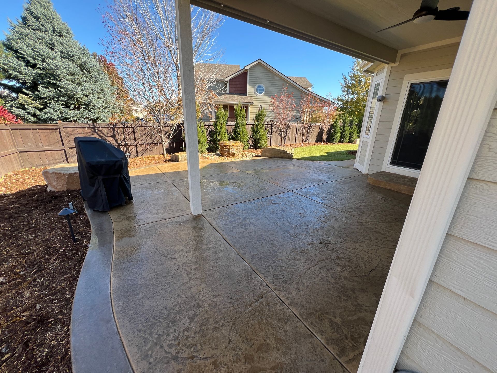 Covered patio with a concrete floor, looking out to a backyard with trees and a house.