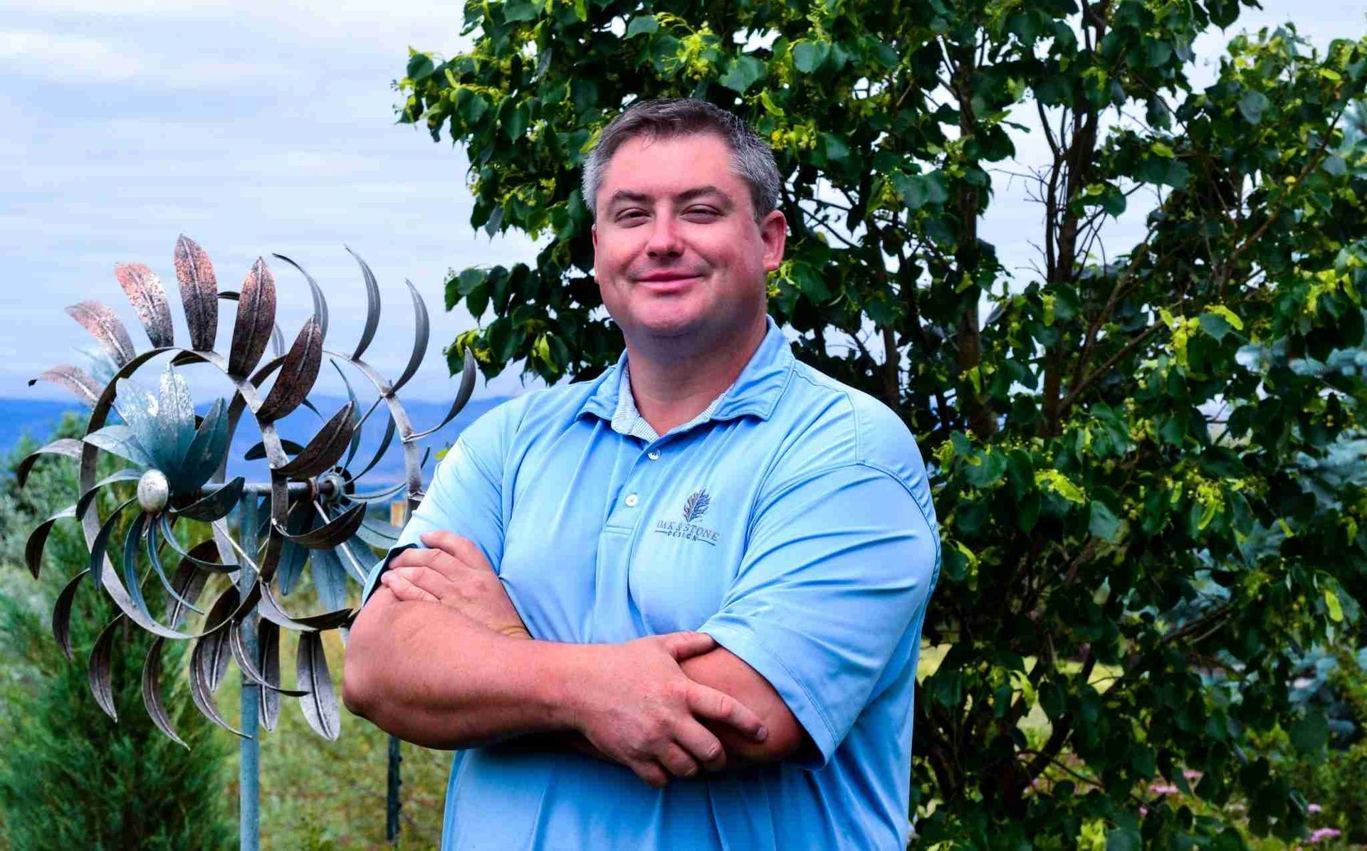 Man in blue shirt, arms crossed, stands in front of a metal wind spinner and greenery, outdoors.