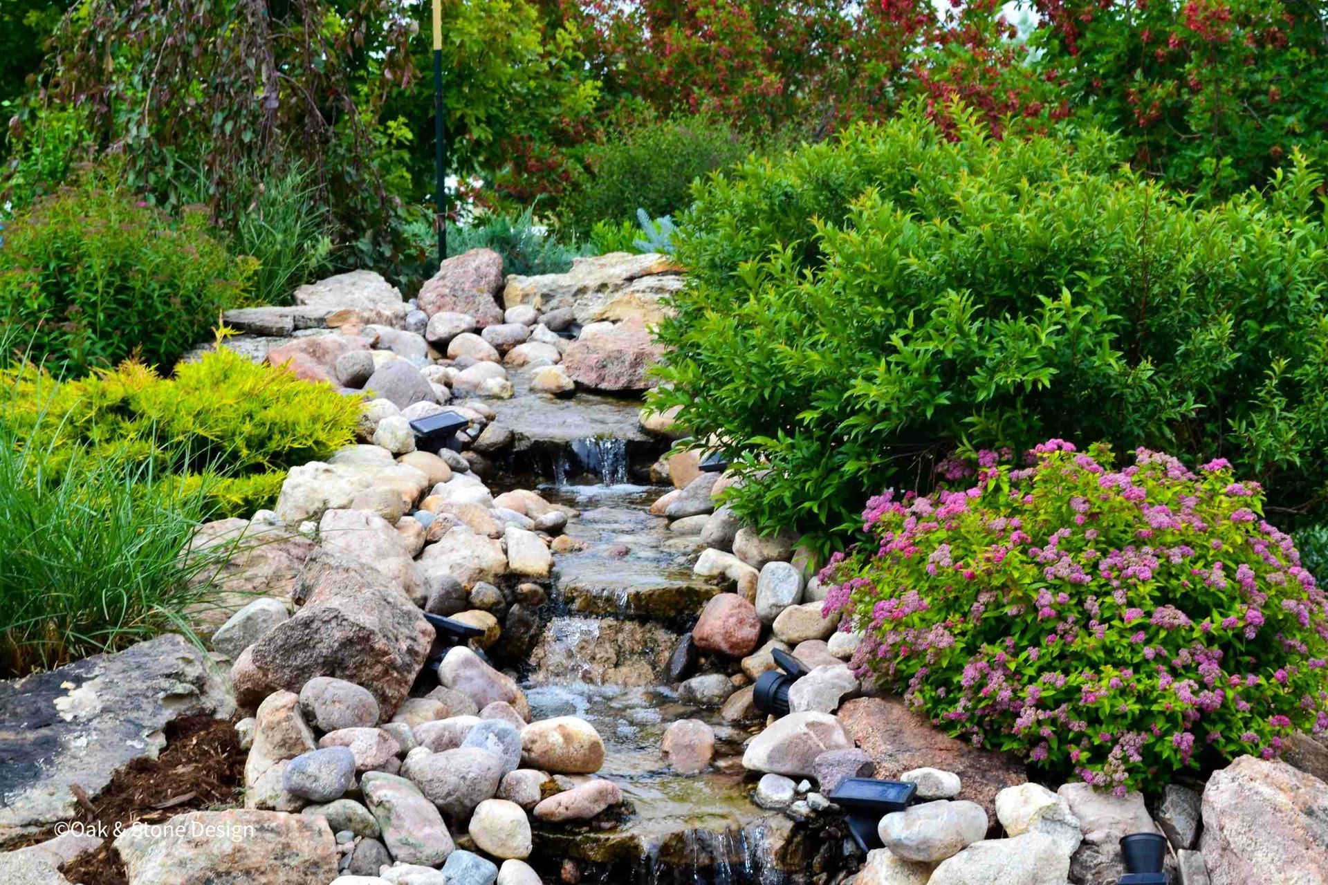 A small waterfall cascading over rocks in a garden, surrounded by green shrubs and pink flowers.