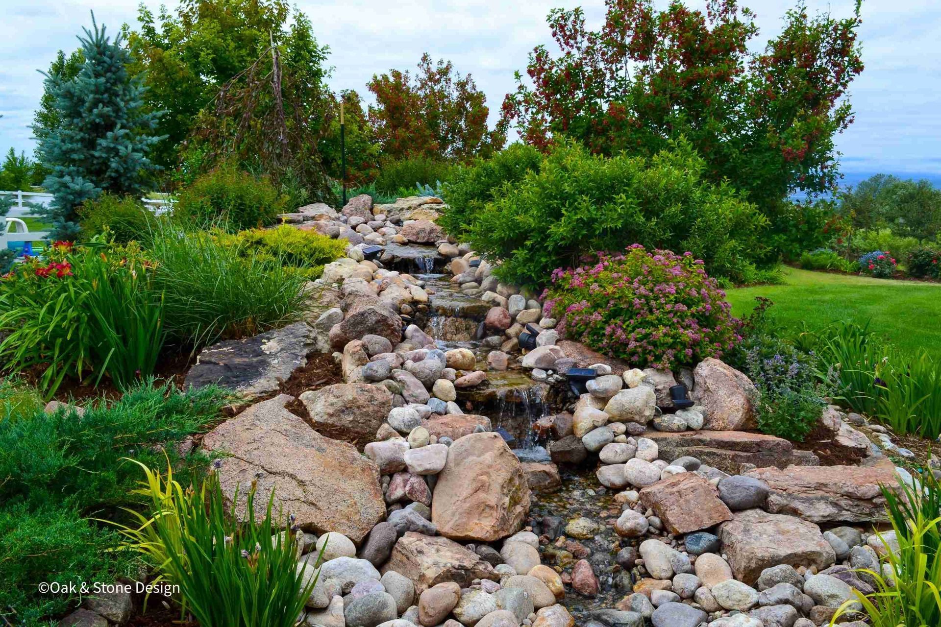 Rocky stream flowing through a landscaped garden, surrounded by green shrubs, trees, and grass.