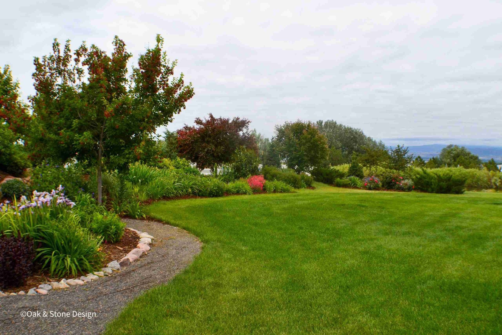 Lush green lawn with a curved gravel path bordered by colorful flowerbeds and trees under a cloudy sky.