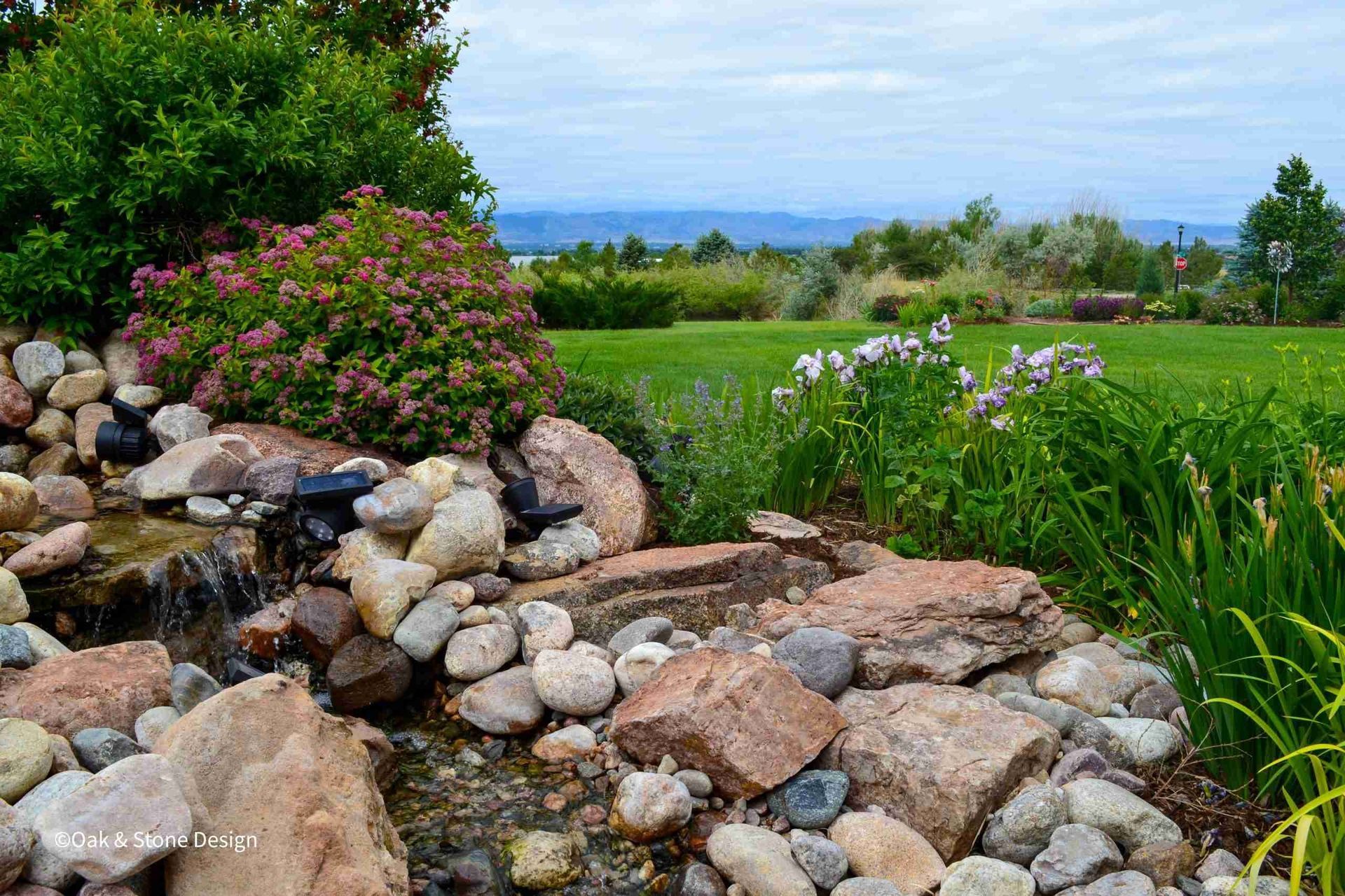 Stone waterfall flowing into a stream, lush green grass, flowering bushes, and distant mountains under a cloudy sky.