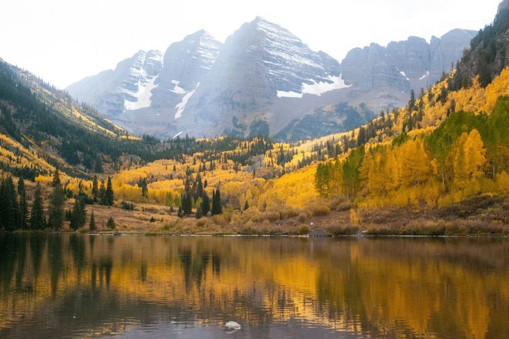 Mountains reflected in a lake, with fall foliage in yellow and green and patches of snow.