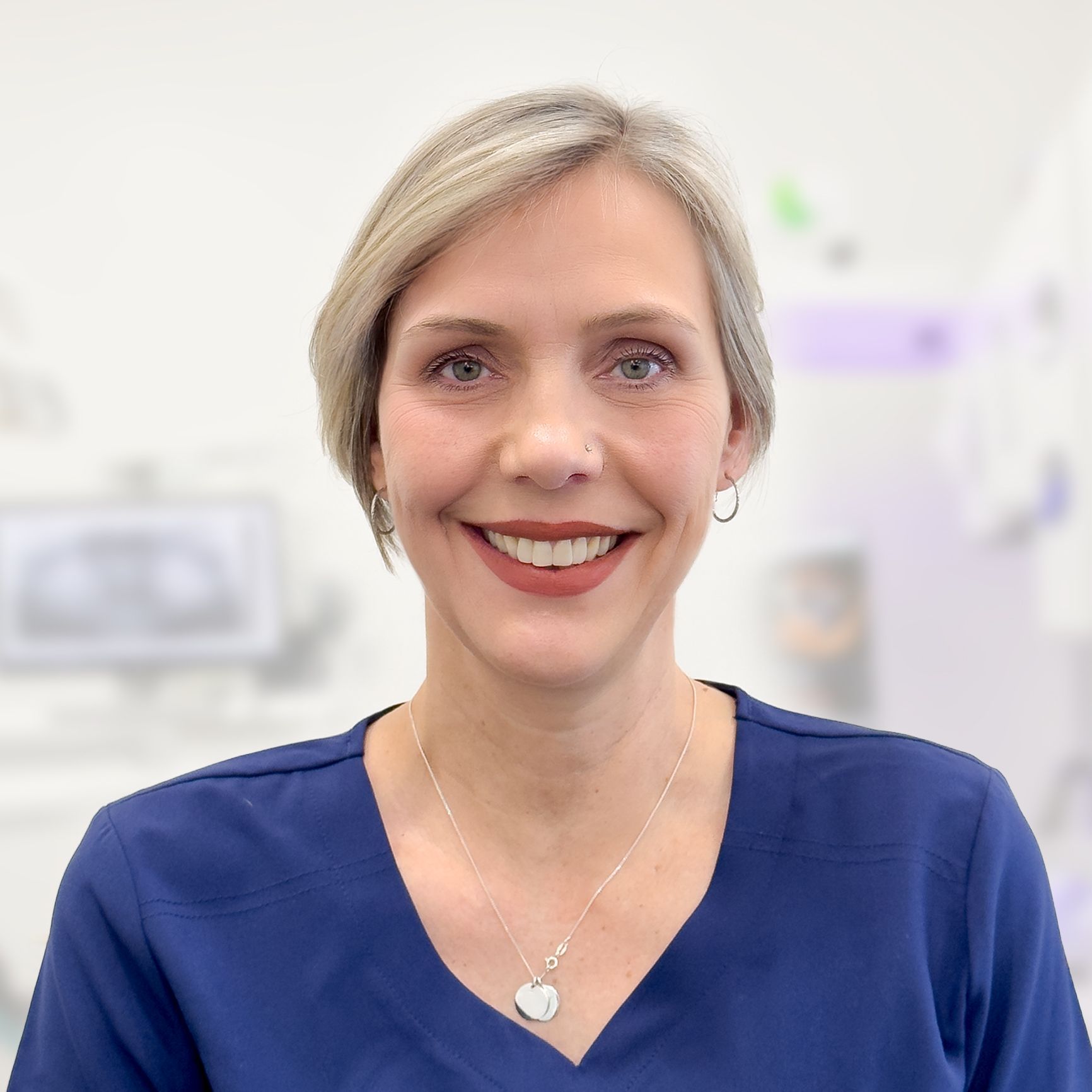 A woman wearing a blue scrub top and a necklace is smiling for the camera.