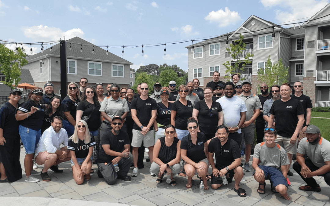 Group of people posing outdoors in front of apartments. Some are kneeling. Sky is blue.