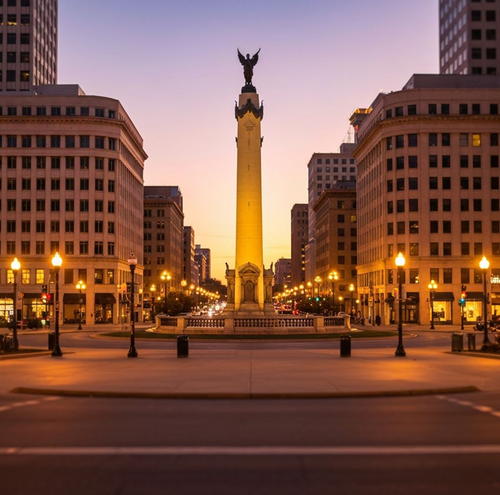 Monument Circle, Indianapolis, at dusk. Tall column with statue, surrounded by buildings and streetlights.