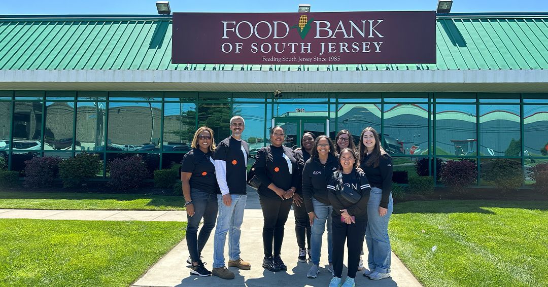 Group stands in front of the Food Bank of South Jersey. They wear black shirts with an orange logo. Building has green roof.