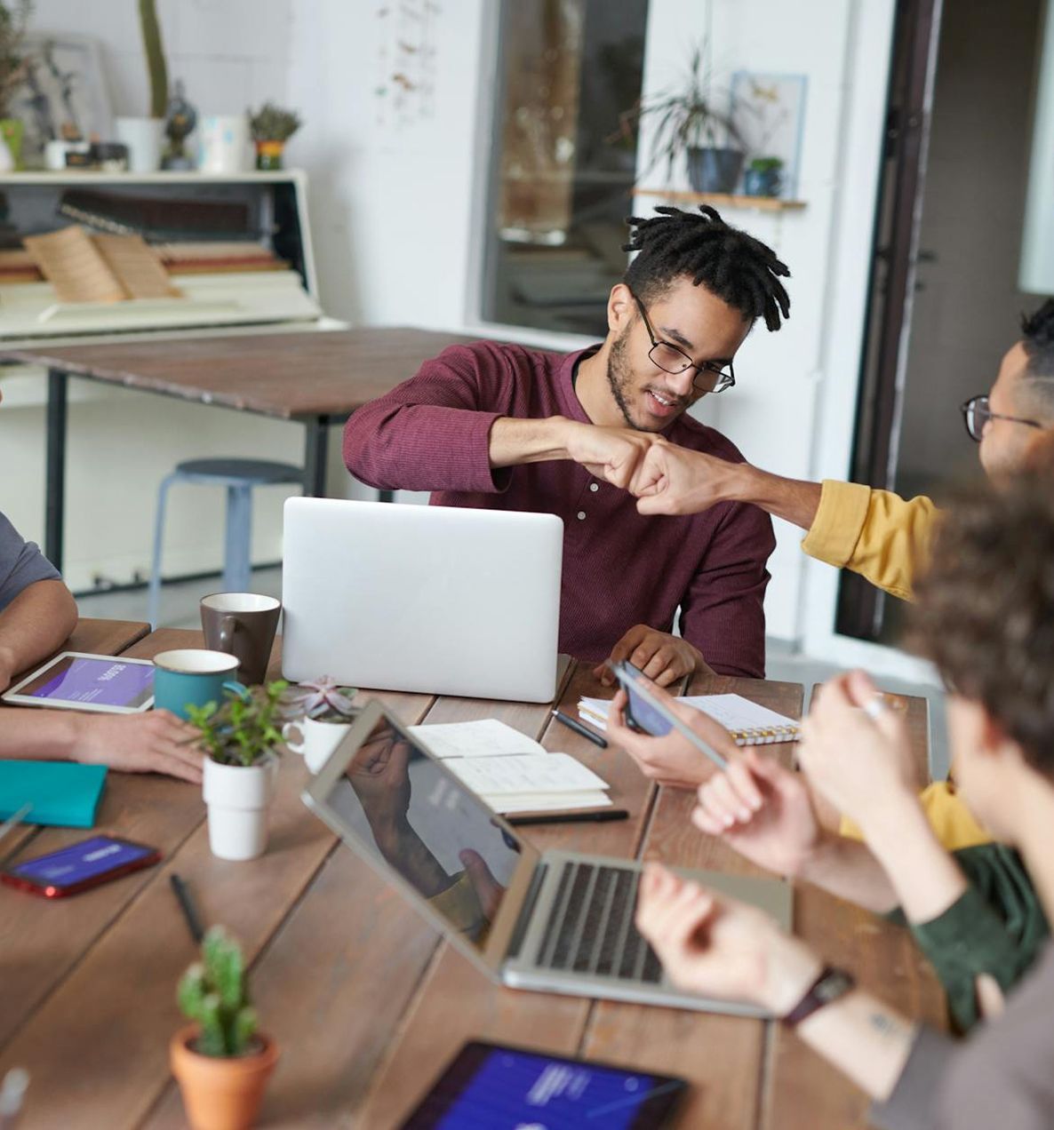 People fist-bumping at a wooden table with laptops, phones, and plants in an office.