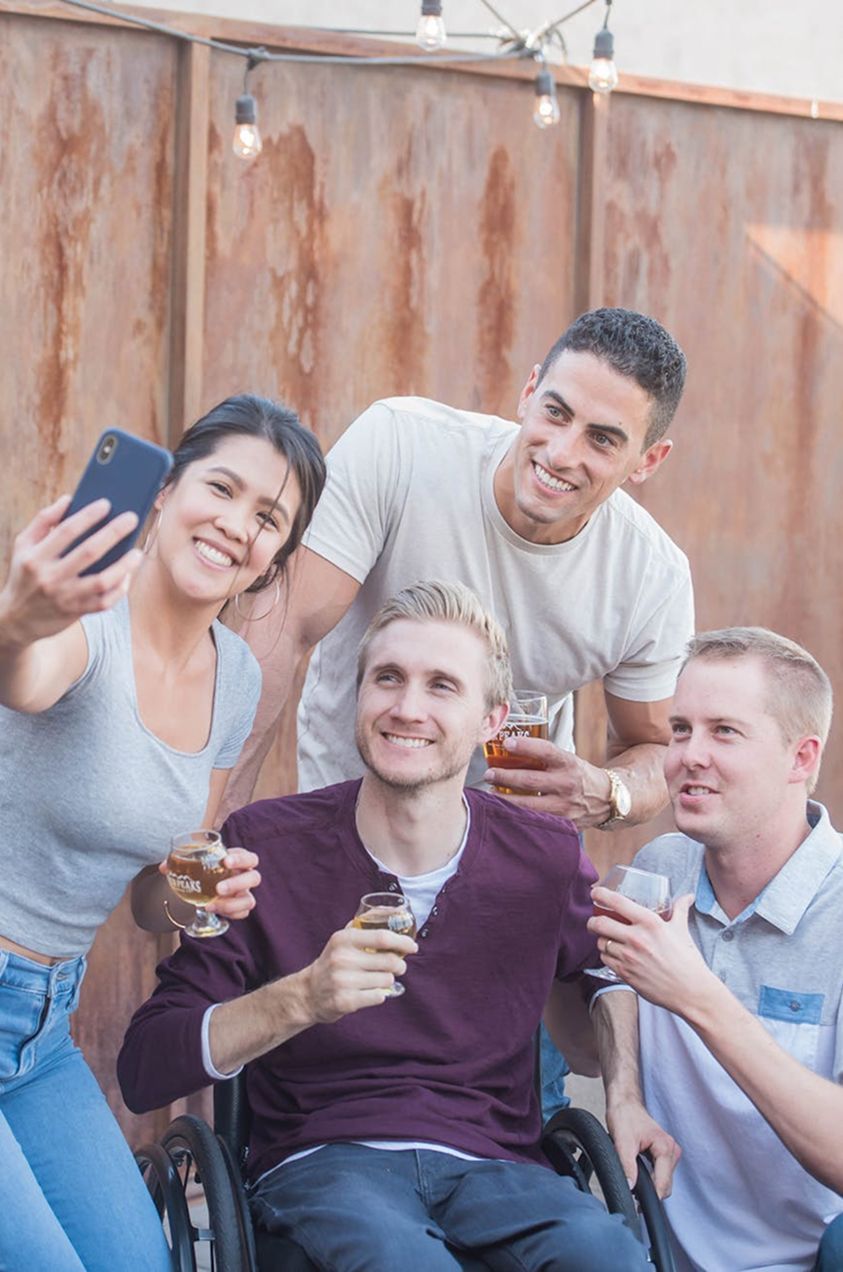 Group of friends taking a selfie, one in a wheelchair, holding drinks outdoors.