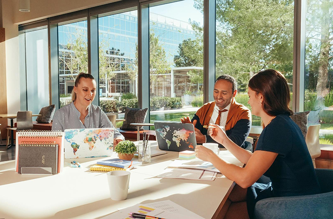 Three people collaborating around a table in a sunlit office, laptops open, smiling, large windows.