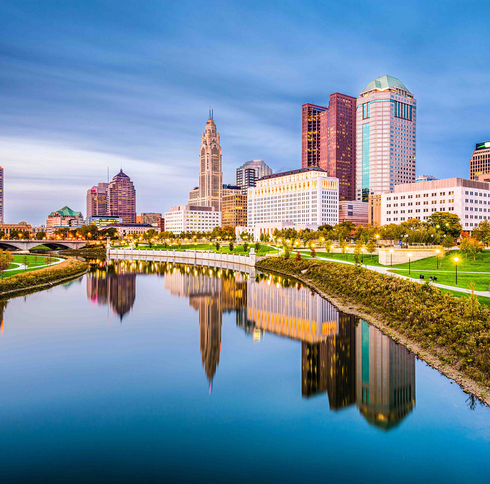 Skyline of Columbus, Ohio, reflected in calm river water; buildings, trees, and blue sky.