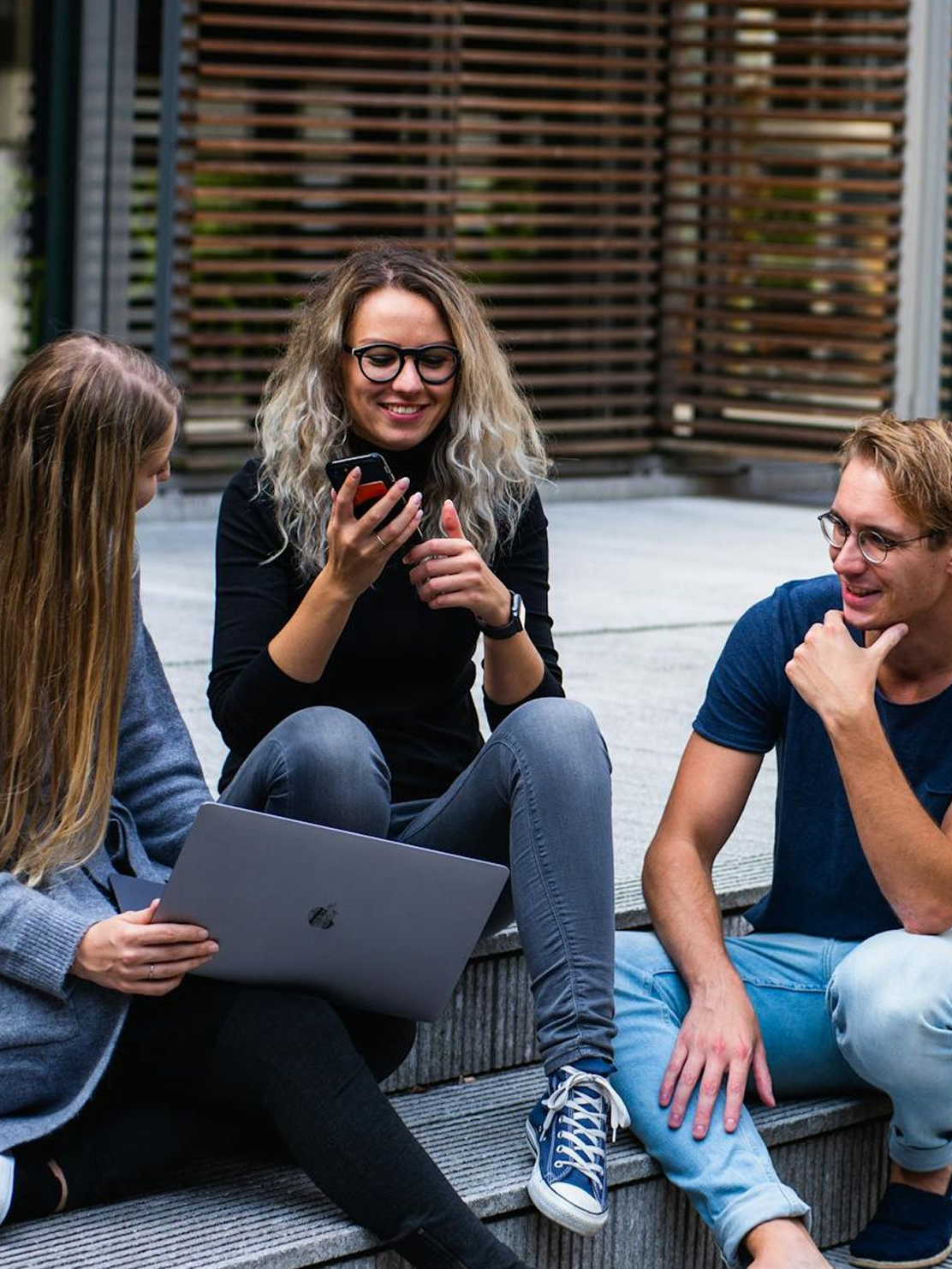 Three people sitting on steps: woman using laptop, woman looking at phone, man smiling.
