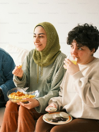 Two people sit, eating snacks and drinking, one wearing a green head covering and sweater, the other with curly hair.