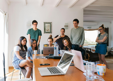 People gathered around laptops at a table, looking at the screens. Indoors, bright room.