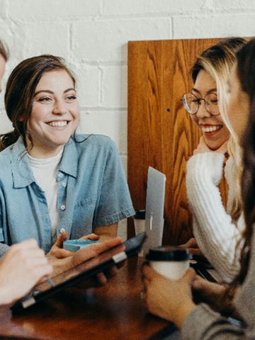 Four people laughing together around a table with a laptop, coffee, and tablet.