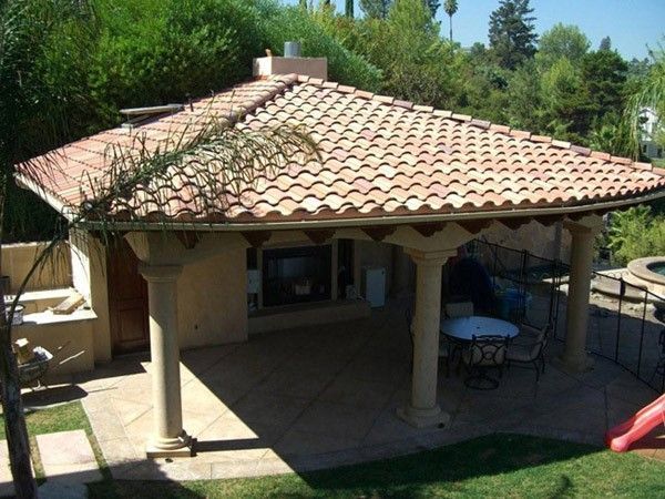 An outdoor pavilion with a tiled roof, pillars, and a fireplace, set in a backyard with greenery and a small play slide.
