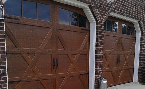 Two brown wooden garage doors with windows, set against a brick wall and framed by white trim.