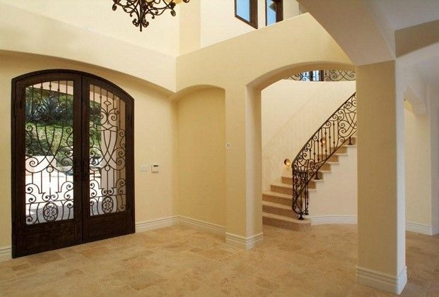 Elegant foyer with double arched doors, ornate ironwork, and a curved staircase. Beige walls and flooring create a spacious feel.