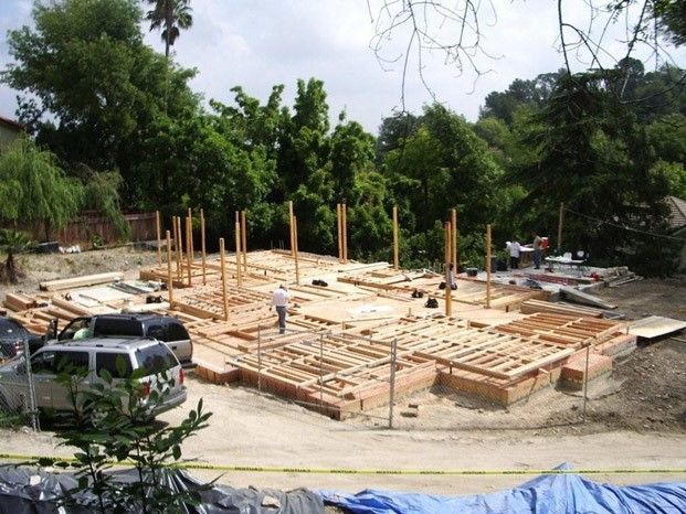 Construction site of a house; wooden framework with workers. Cars and trees surround the work area.