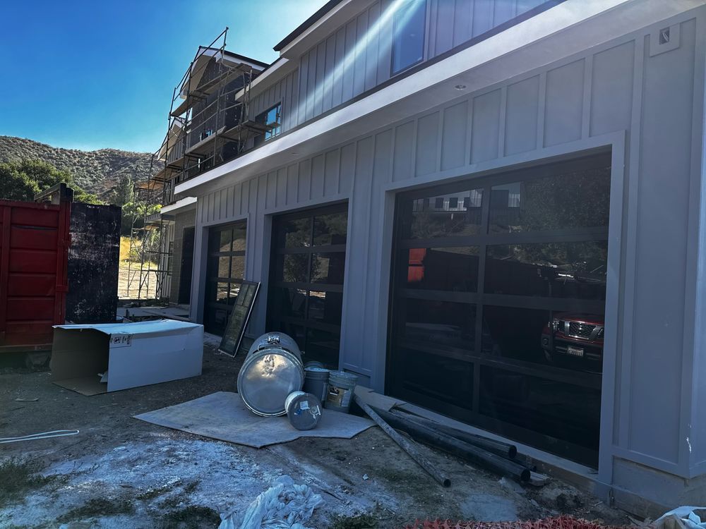 Exterior of a light gray house with three garage doors, reflecting a car inside. Construction materials and scaffolding are visible.