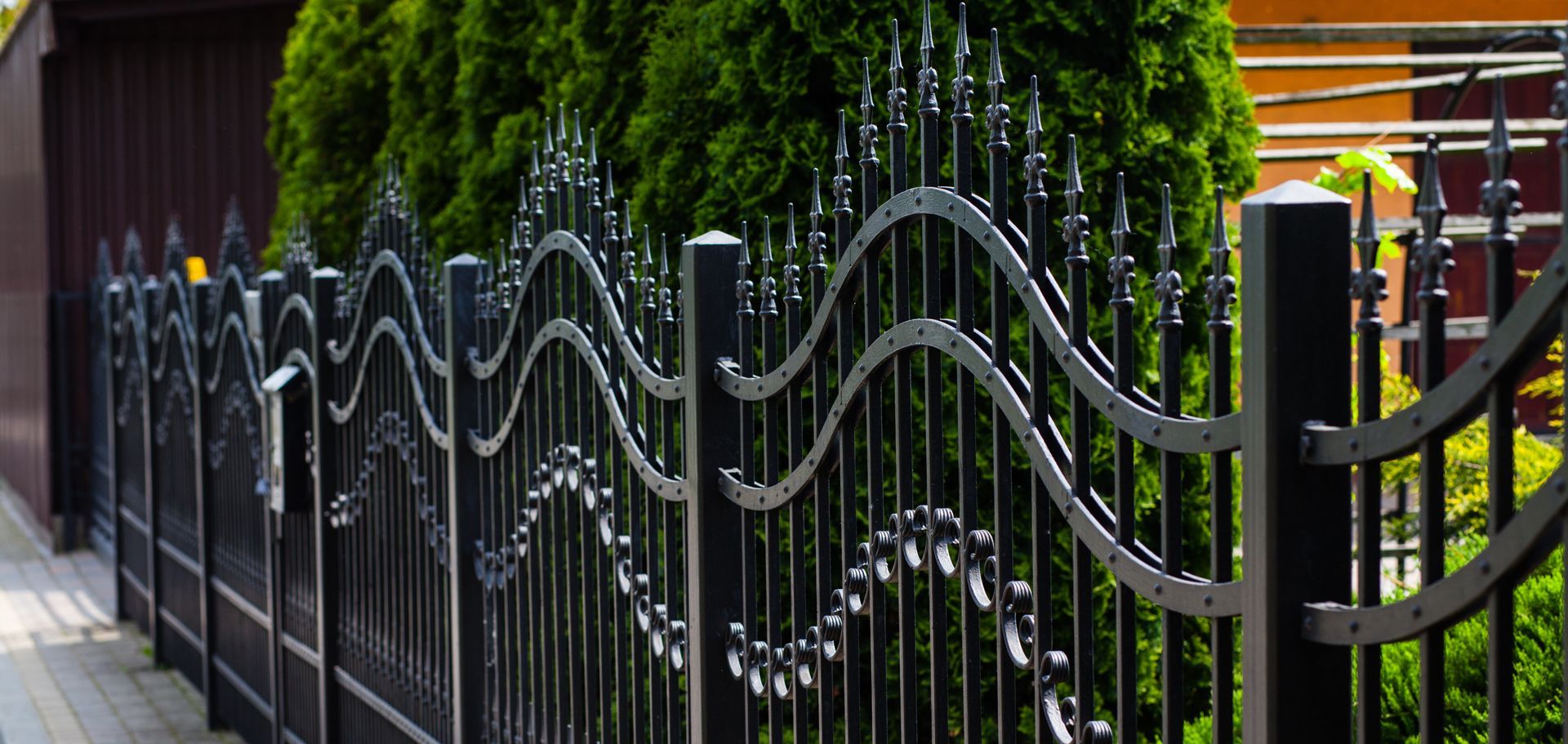 Black wrought iron fence with decorative wavy design, set against green foliage and a brown building.