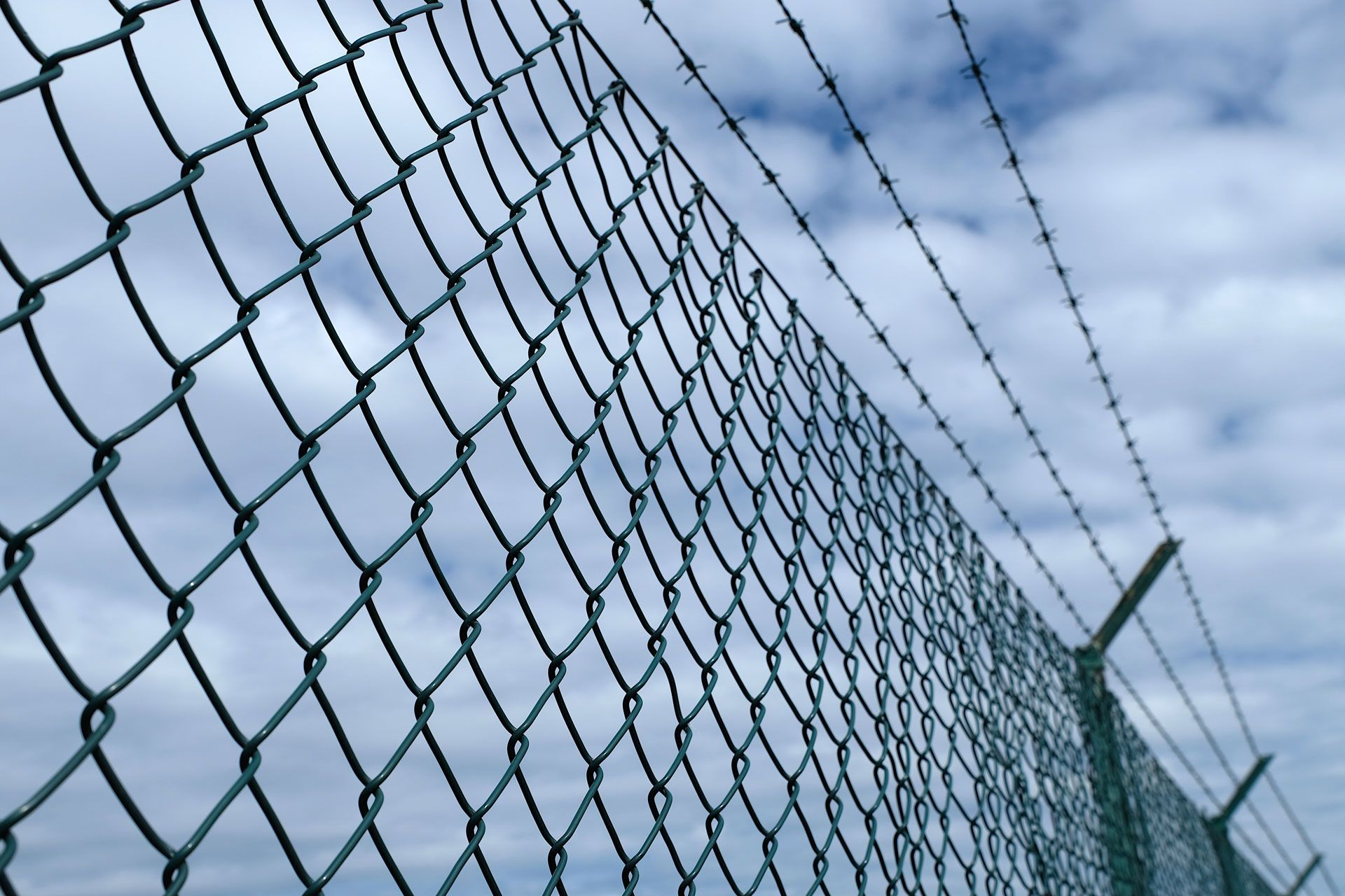 Chain-link fence with barbed wire against a cloudy sky.
