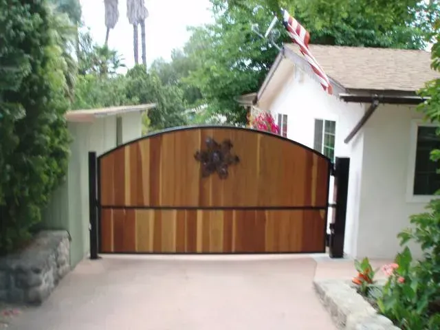 Wooden arched gate with black frame, metal flower ornament.  Facing small white house, driveway.