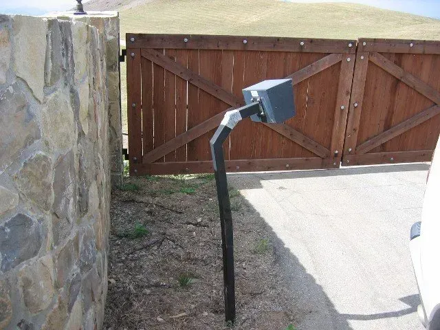 Black security camera on a curved metal pole next to a brown wooden gate and stone wall.