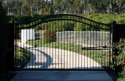Black metal driveway gate on concrete, with curved top, opening to a curved driveway and greenery.