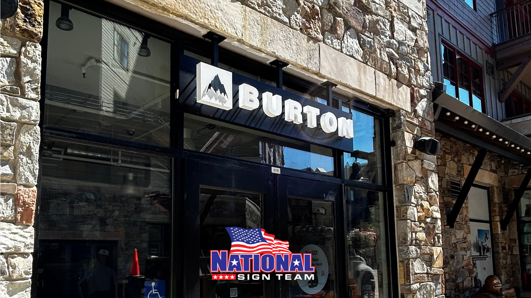 Burton storefront with sign and window display, stone building facade, US flag.
