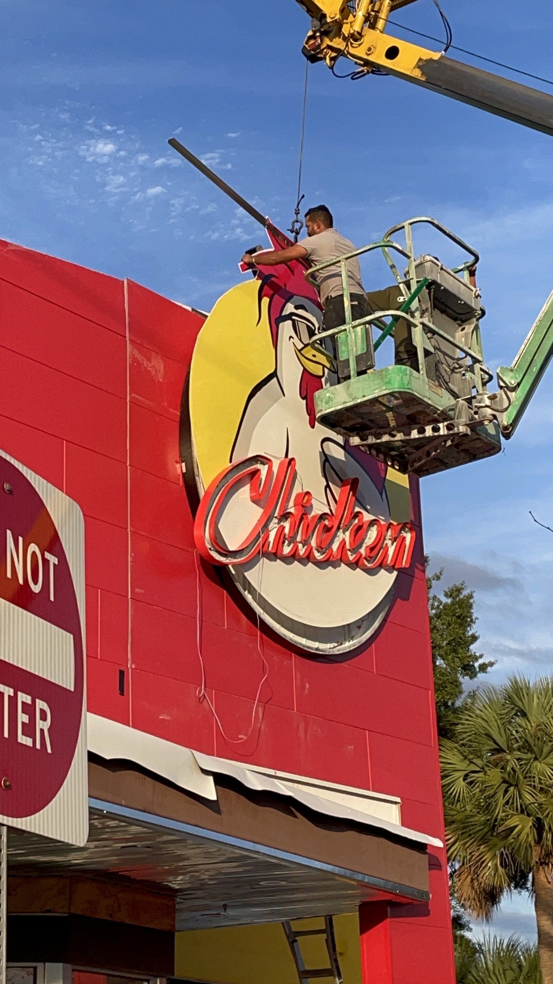 A man in a crane is painting a chicken sign on the side of a building.