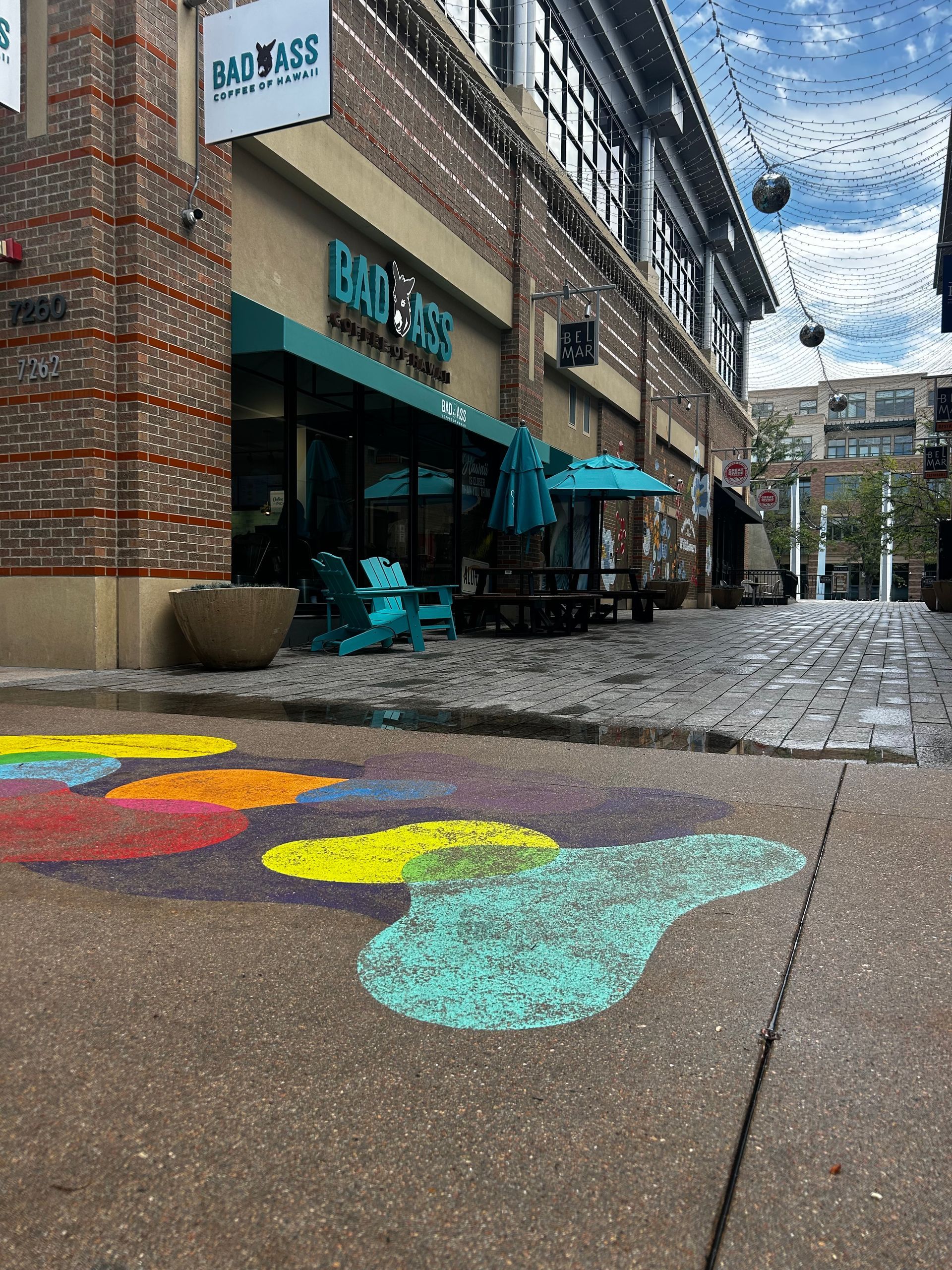Colorful sidewalk art outside a café with turquoise chairs and umbrellas on a cobblestone street.