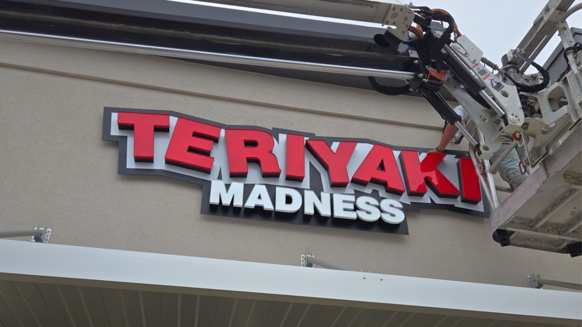 Teriyaki Madness sign being worked on by a lift; red and white letters on tan building.