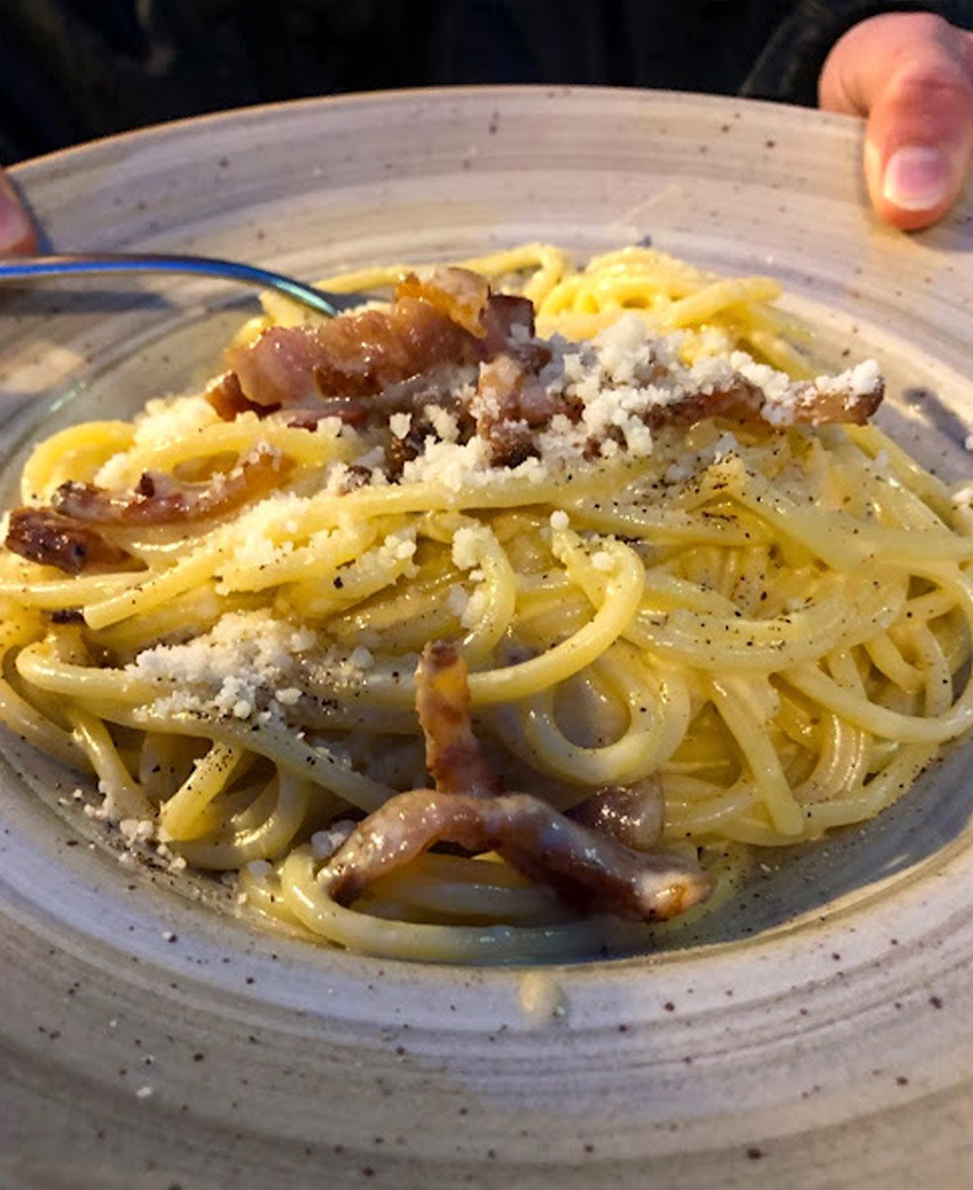 Una persona sta mangiando spaghetti con pancetta e formaggio su un piatto con una forchetta.