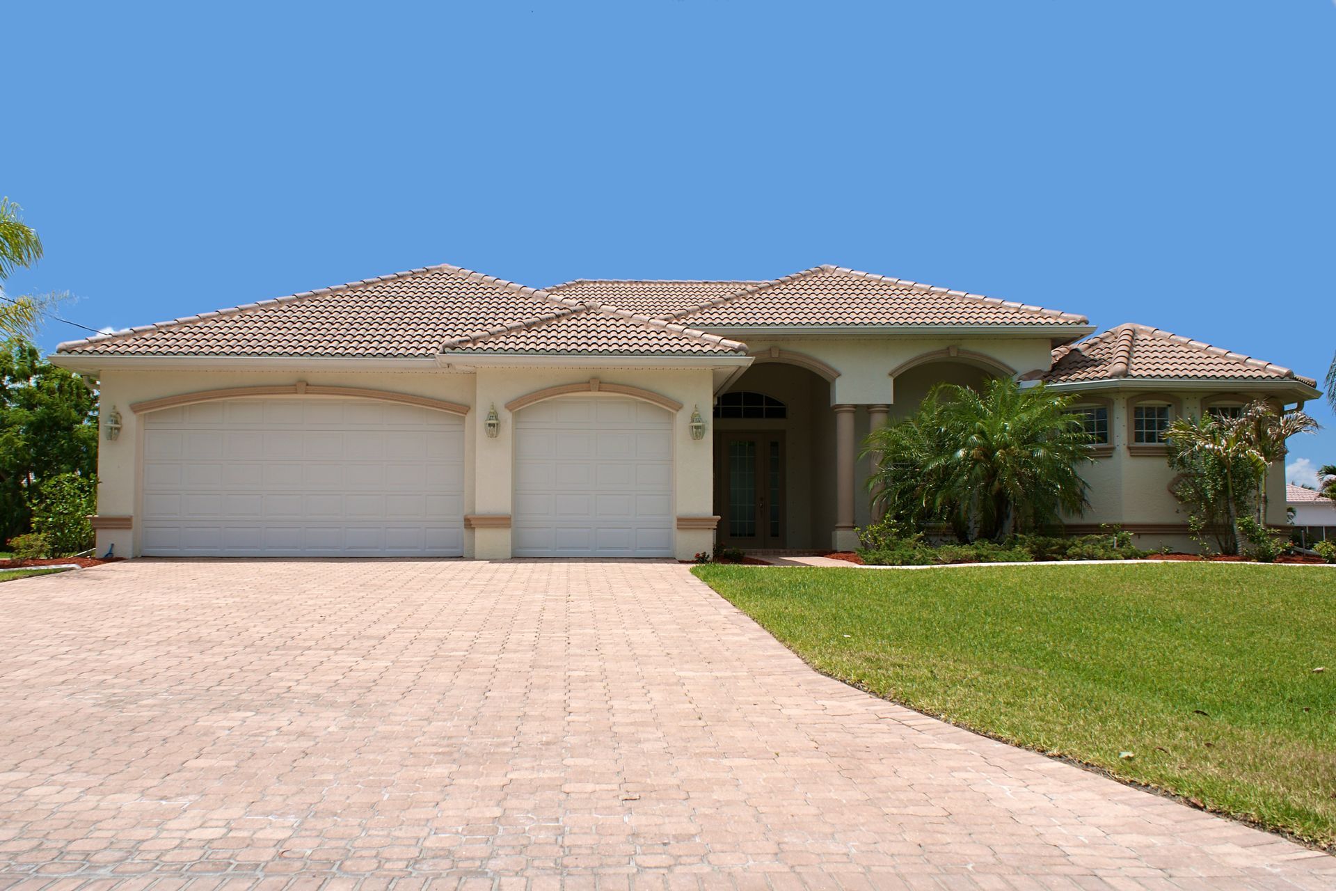 A beige, single-story house with a tile roof, two-car garage, and a paved driveway. It sits on a green lawn under a blue sky.