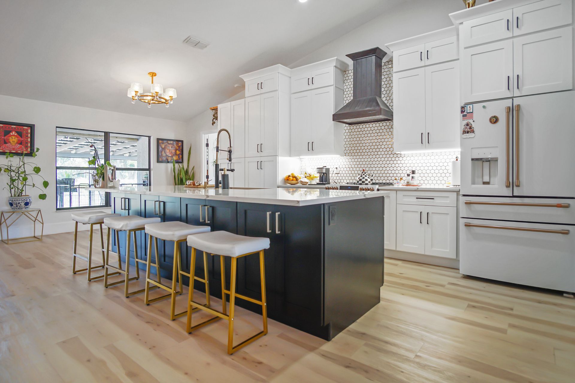 Modern kitchen with white cabinets, black island, and gold accents. Blond wood flooring, stainless steel appliances, and a chandelier.