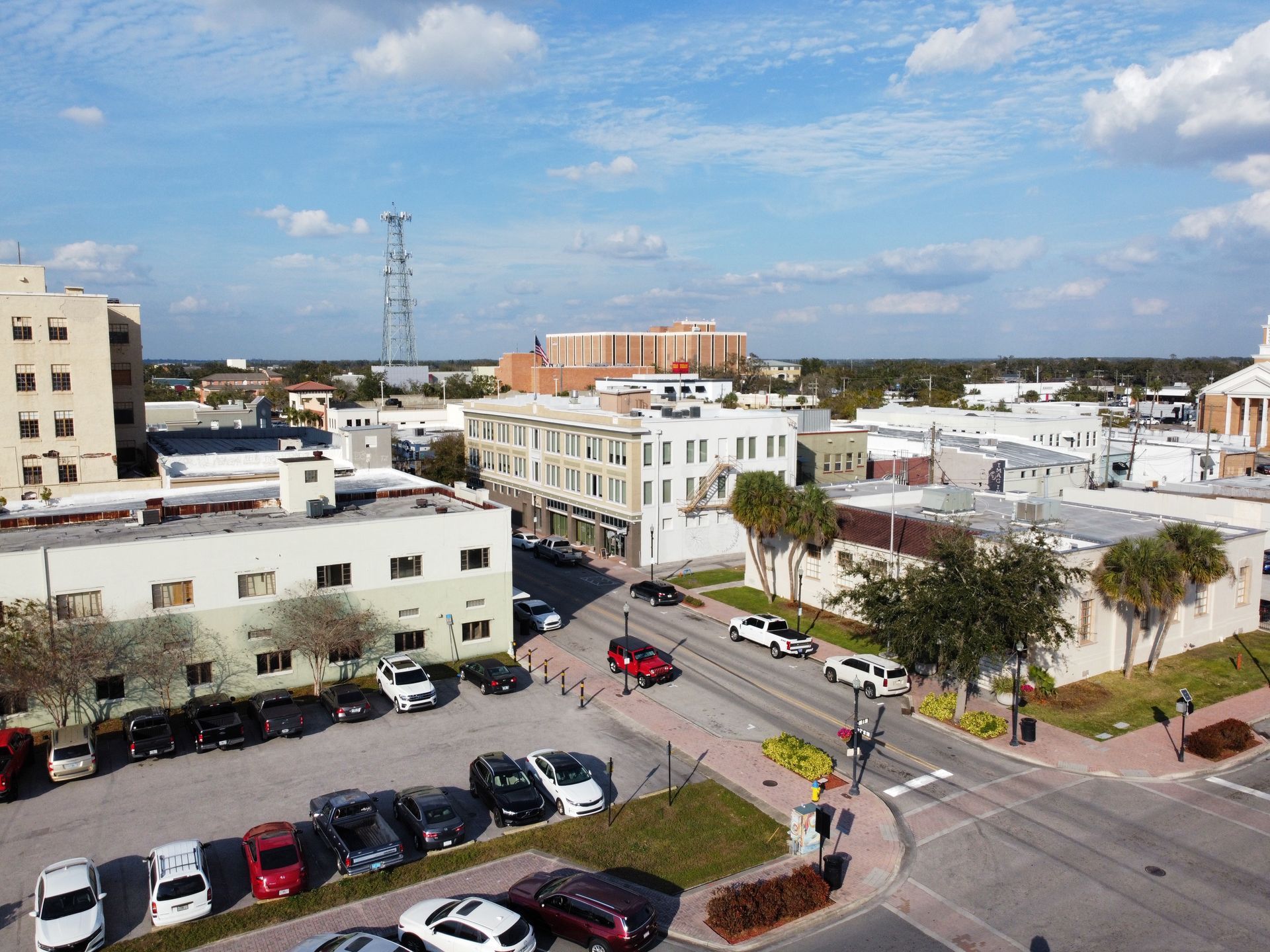 Aerial view of a city street with parked cars, buildings, and a tall communication tower under a blue sky with clouds.