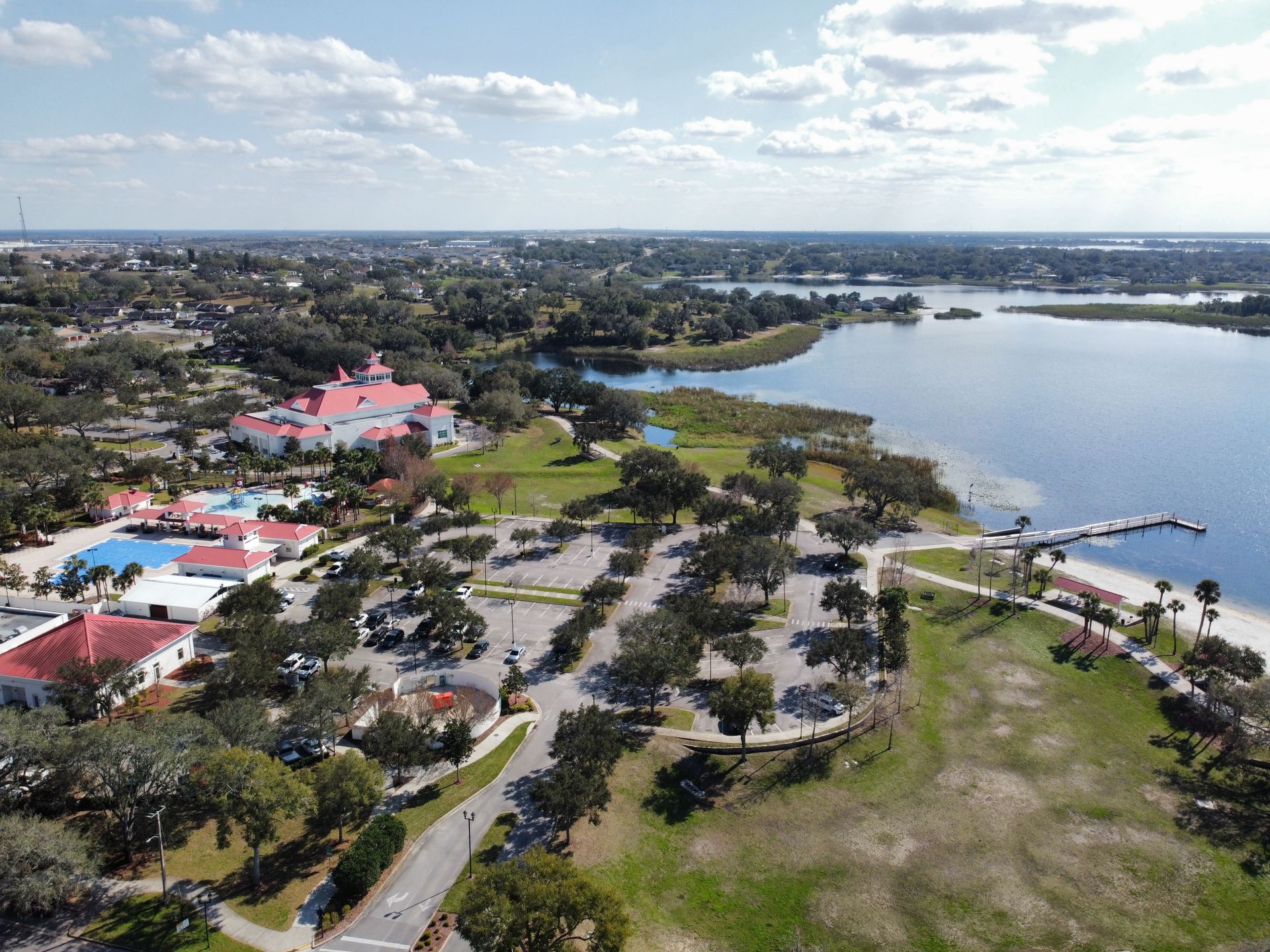 Aerial view of a park and lakeside area. A large white building with red roofs is on the left, and a small beach and pier are on the right.