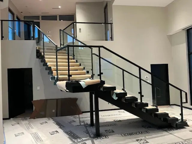 Modern staircase with black metal frames, glass railings, and light-colored treads in an unfinished construction space.