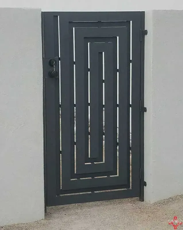 A dark grey, rectangular metal pedestrian gate with a modern, geometric spiral cutout design, set between two white walls.