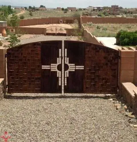 Brown metal gate with white geometric design at the end of a gravel driveway, with a wall and buildings beyond