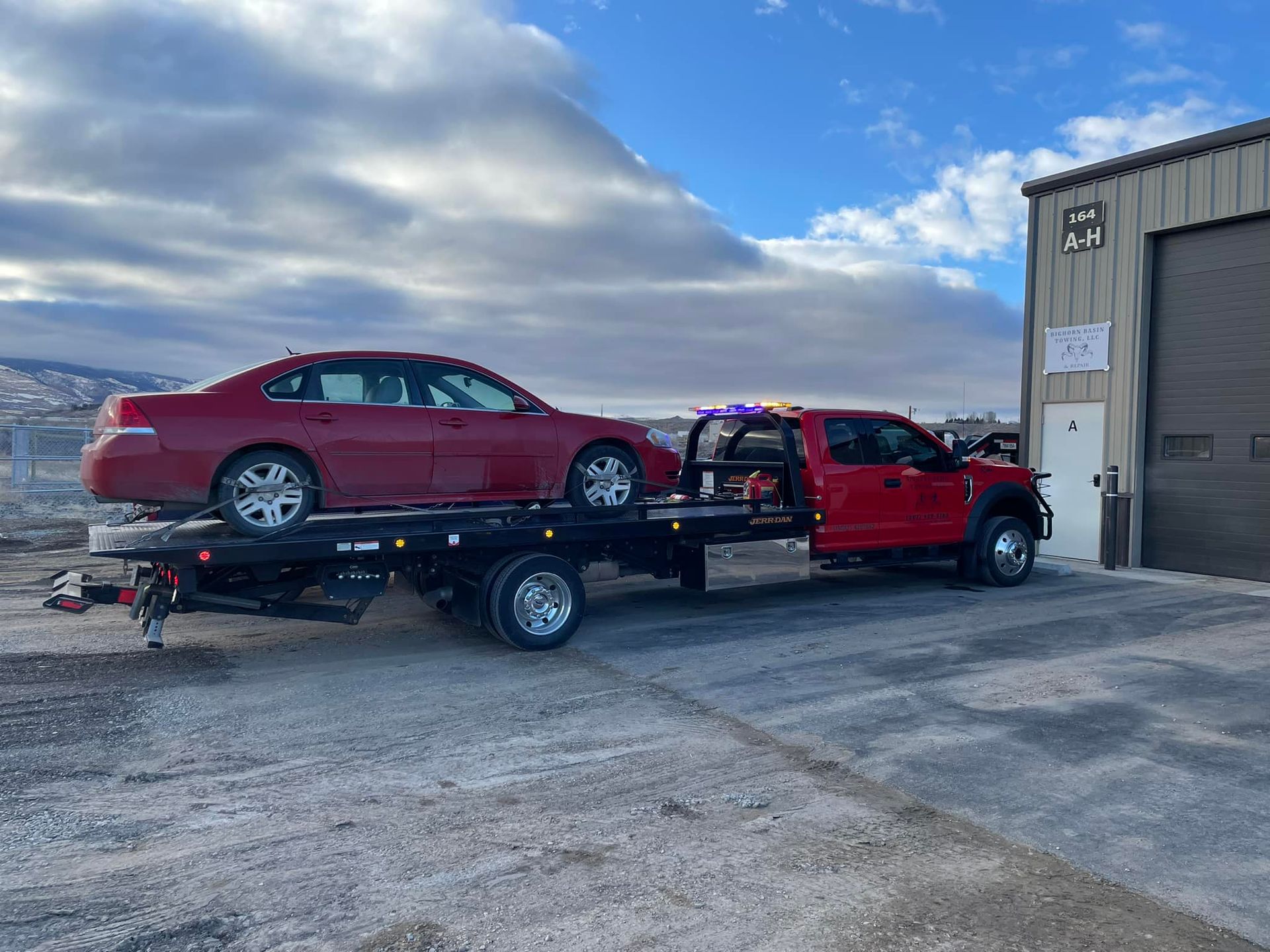 A red tow truck with a red car on its flatbed is parked near a building on a cloudy day.