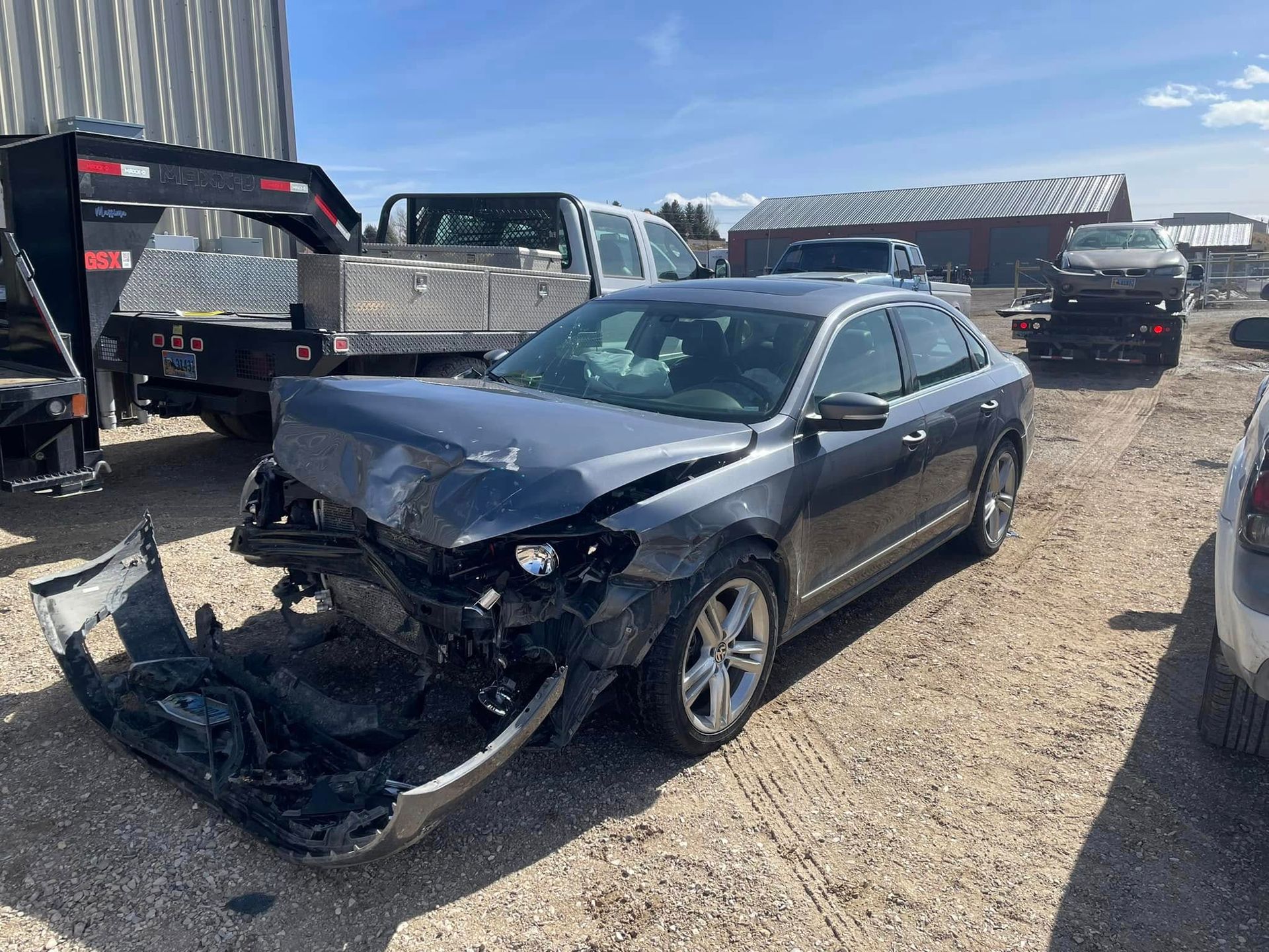 A damaged grey sedan in a salvage yard with front-end collision damage.