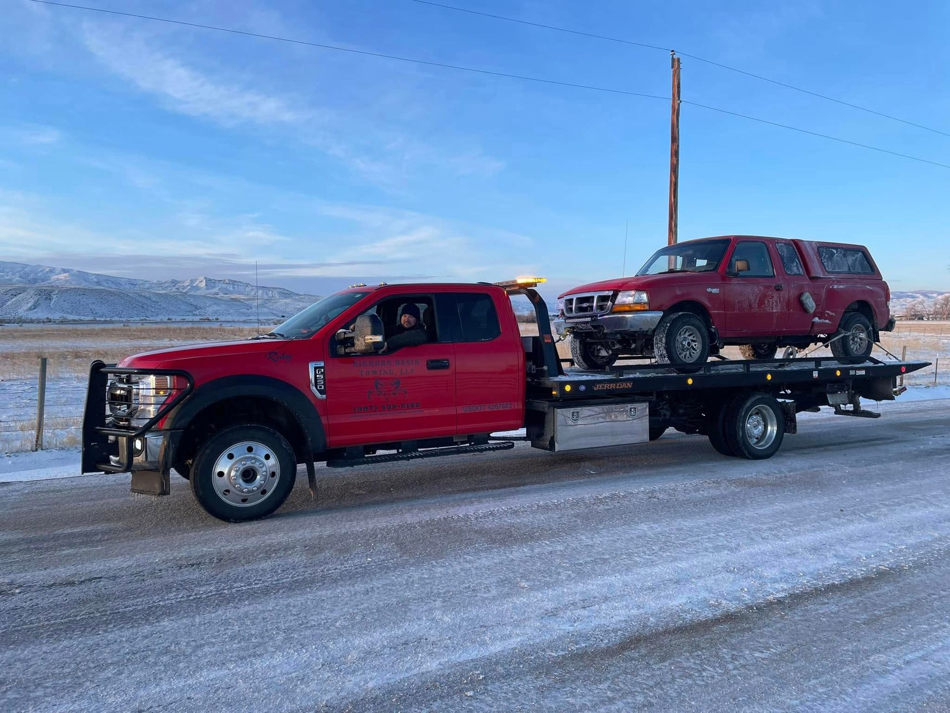 Red tow truck transporting a red pickup truck on a snowy road in a rural setting.