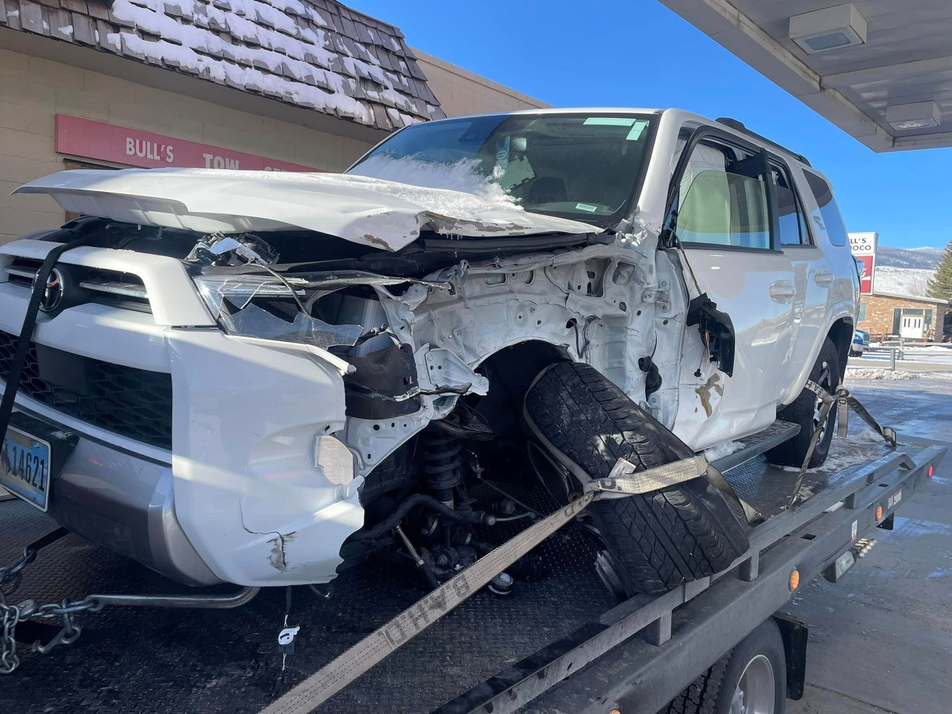A severely damaged white SUV on a tow truck at a gas station. The front end is heavily crumpled.