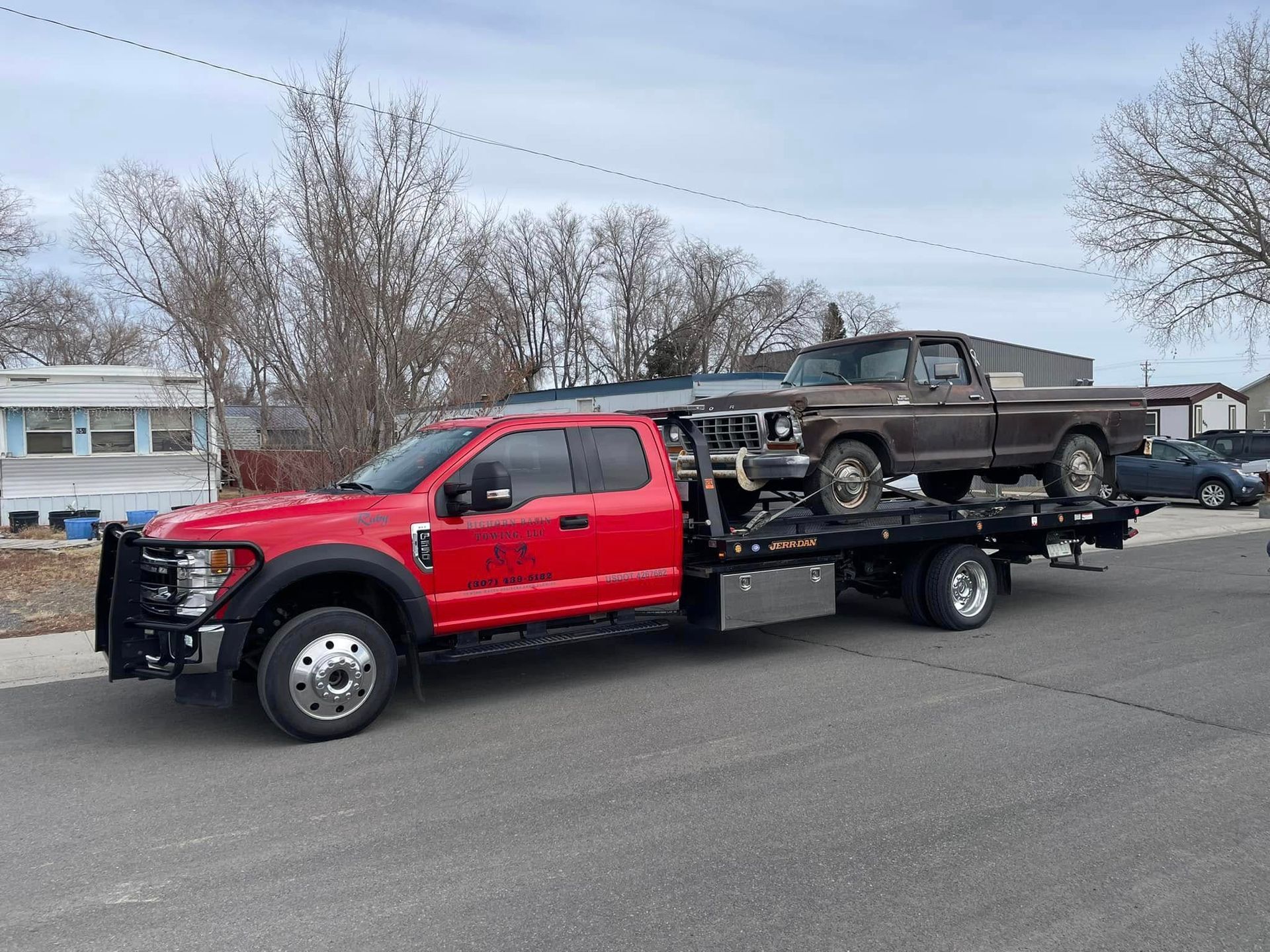 Red tow truck carrying a brown pickup truck on a flatbed trailer on a street.