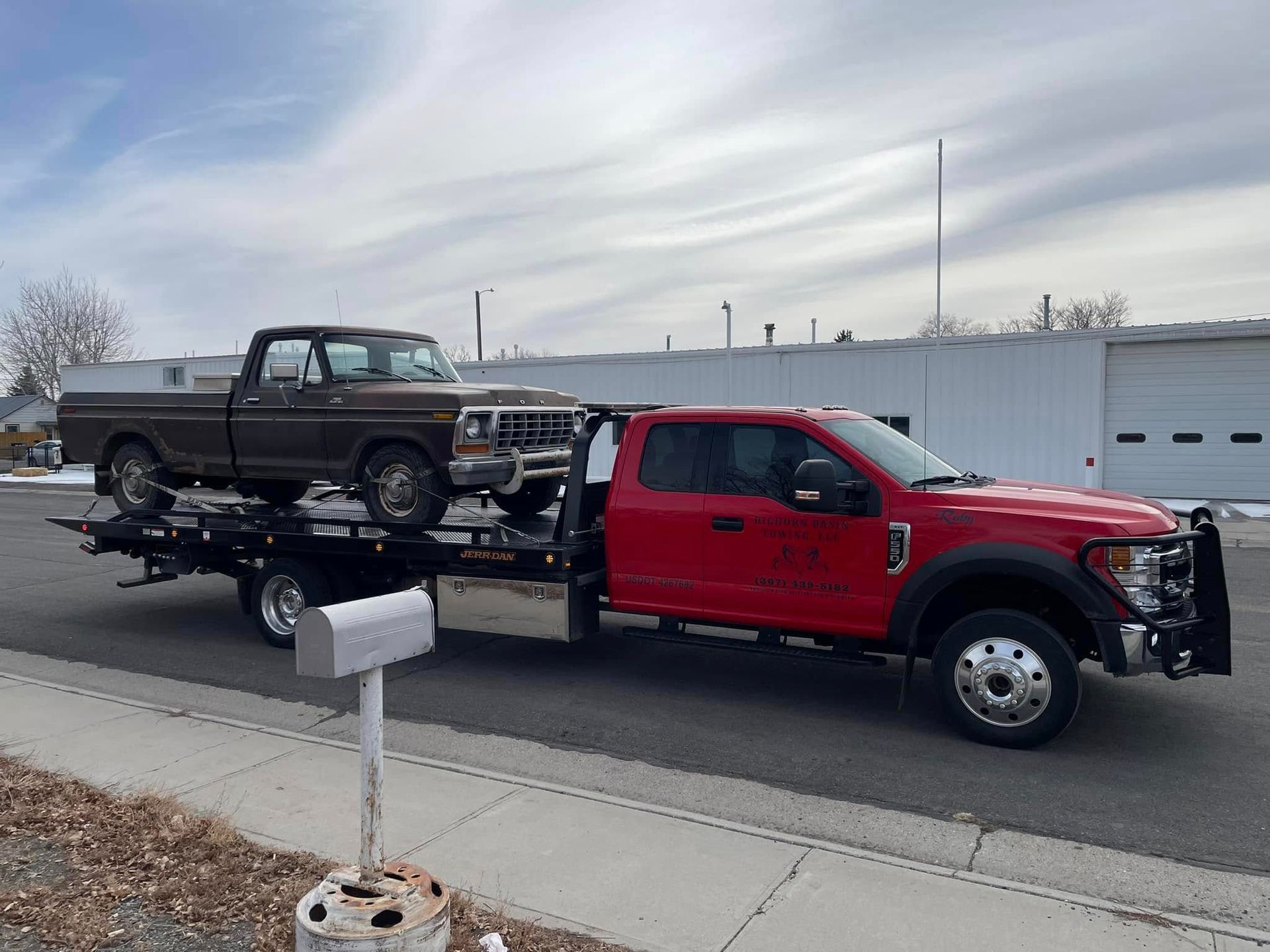 A brown vintage pickup truck loaded on a red tow truck on a street in front of a building.