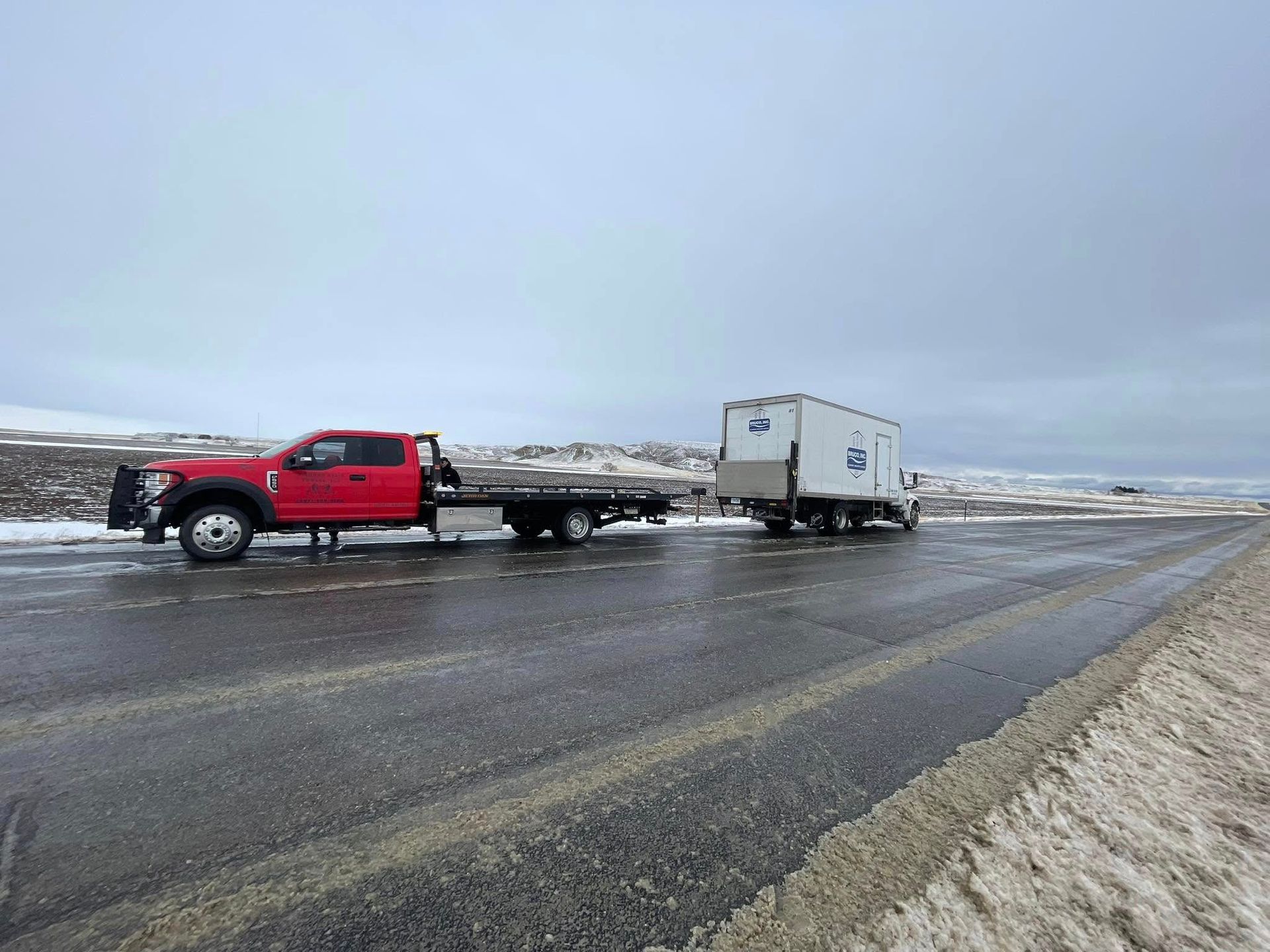 Red tow truck pulling a white mobile building on a snow-covered road under an overcast sky.